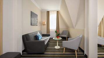 Una habitación de hotel moderna con un sofá negro, un sillón gris, una mesita redonda con un jarrón de flores y una alfombra a rayas.