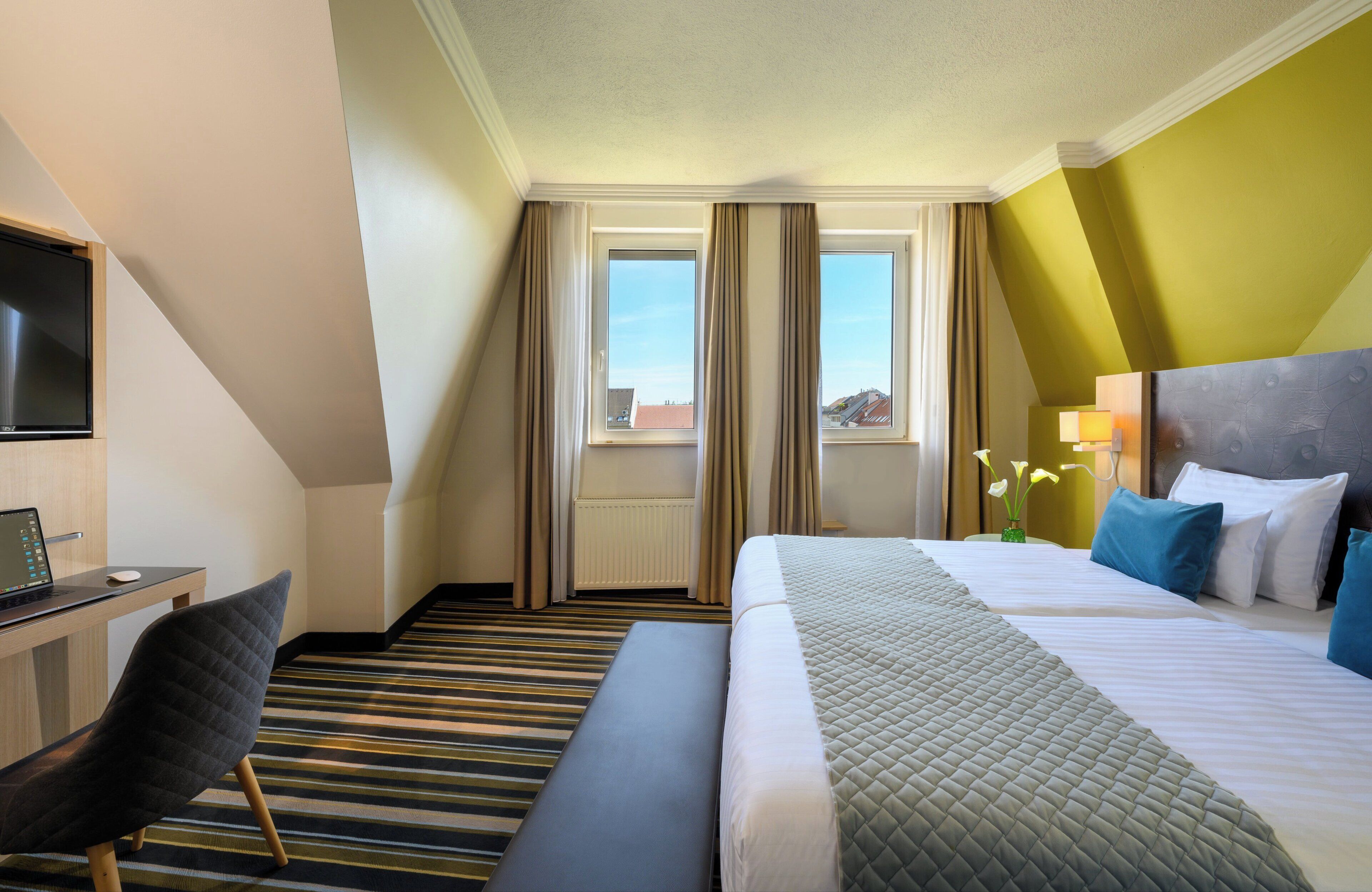 A hotel room with a bed, a desk with a laptop, a chair, a bench, a TV, and a window with curtains.