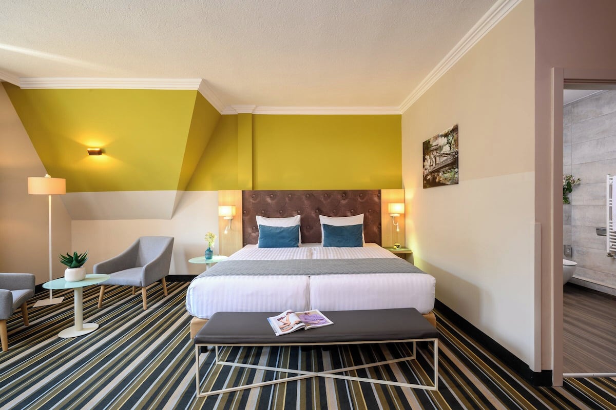 A hotel room with a large bed, a small table with a plant, a chair, and a bench.