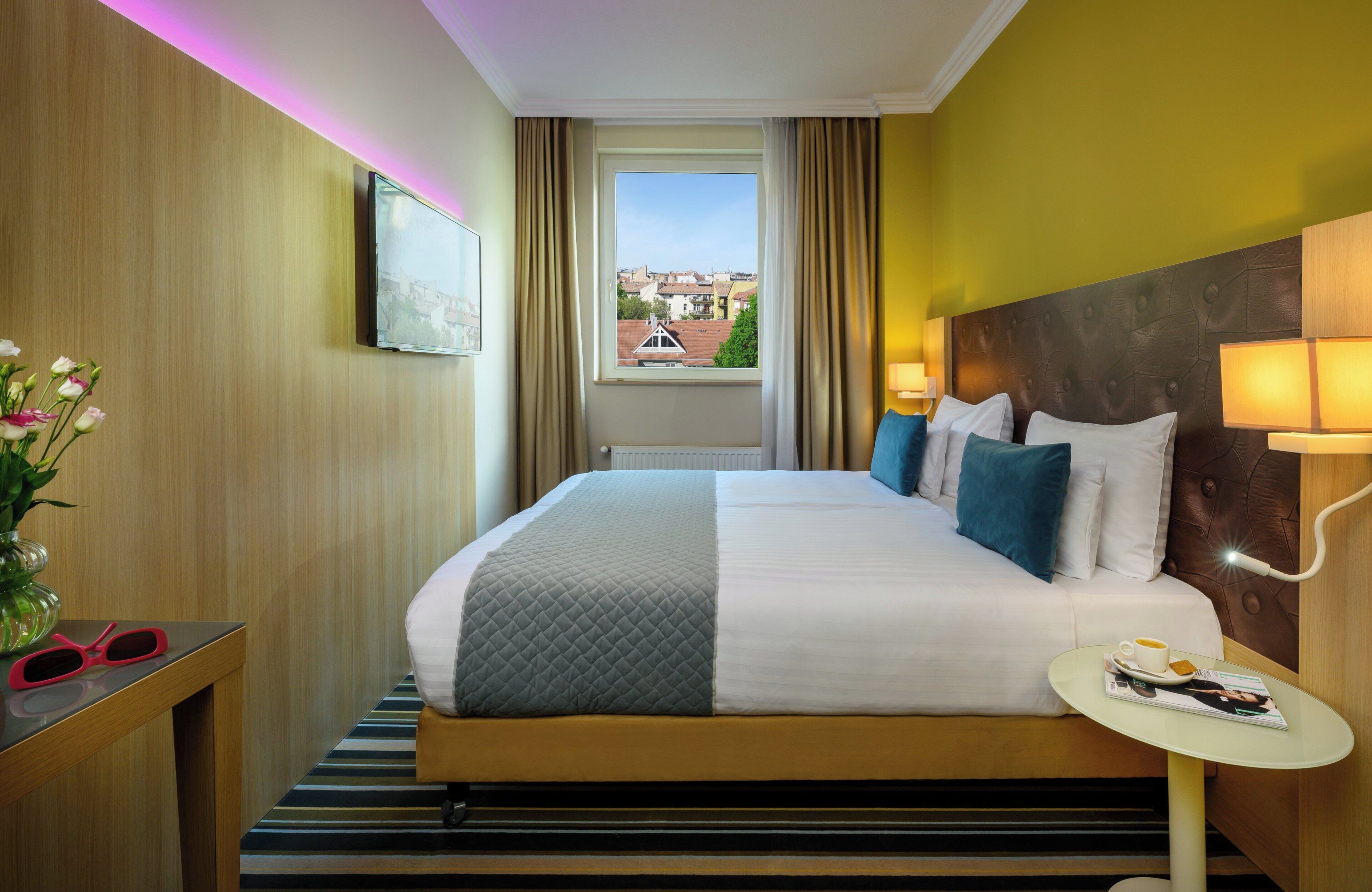 Ein Hotelzimmer mit einem großen Bett, einem Nachttisch mit Lampe, einem Fenster mit Vorhängen und Blick auf Gebäude.