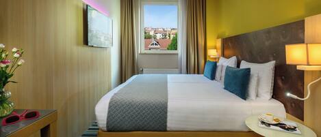Ein Hotelzimmer mit einem großen Bett, einem Nachttisch mit Lampe, einem Fenster mit Vorhängen und Blick auf Gebäude.