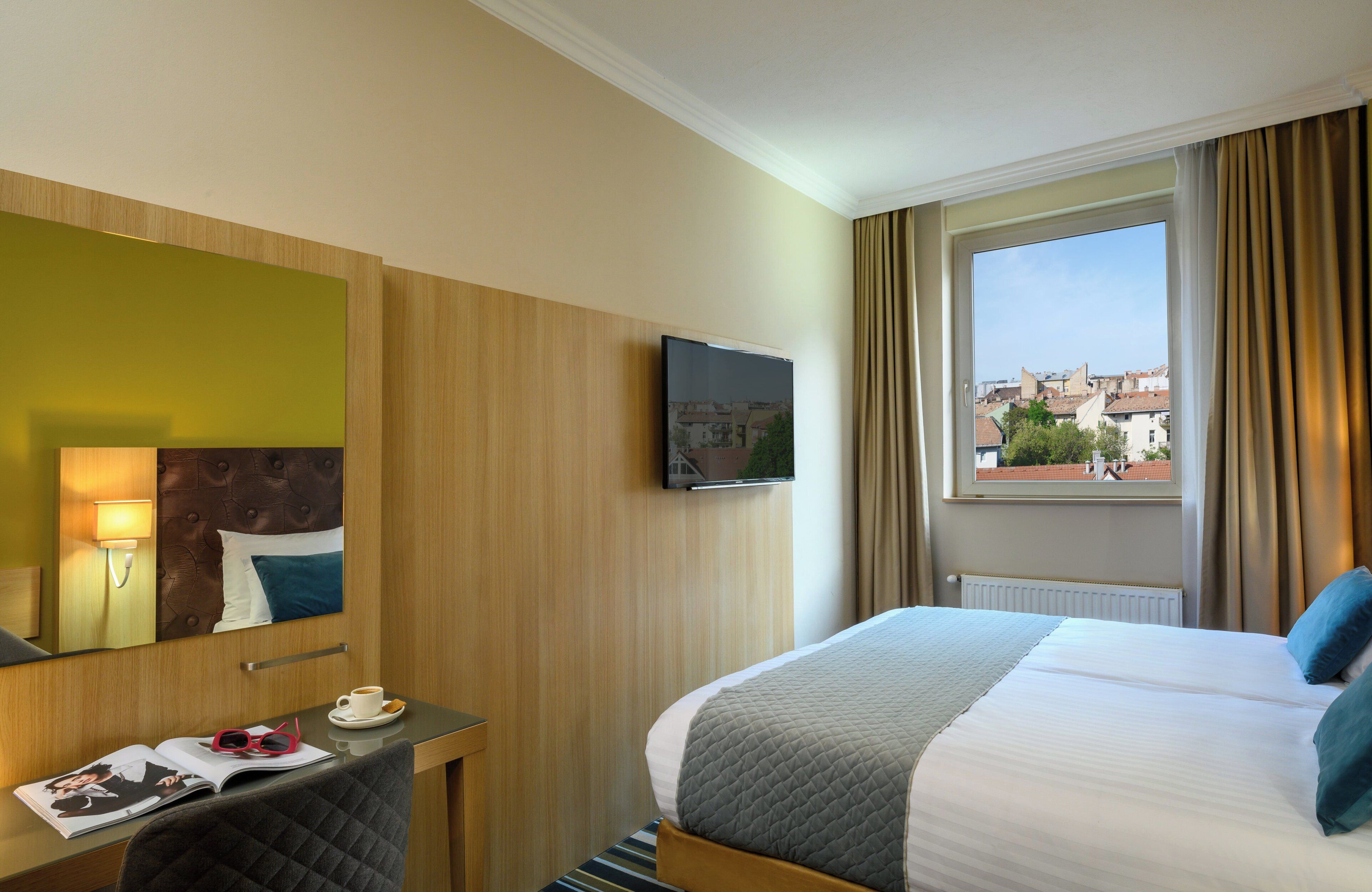 Quarto de hotel com uma cama, televisão, janela com cortinas, escrivaninha com uma chávena e revista, e vista para edifícios.