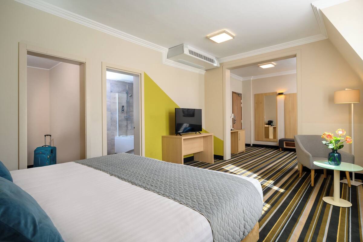 A hotel room with a large bed, a desk with a TV, a chair, a small table with flowers, and a suitcase.