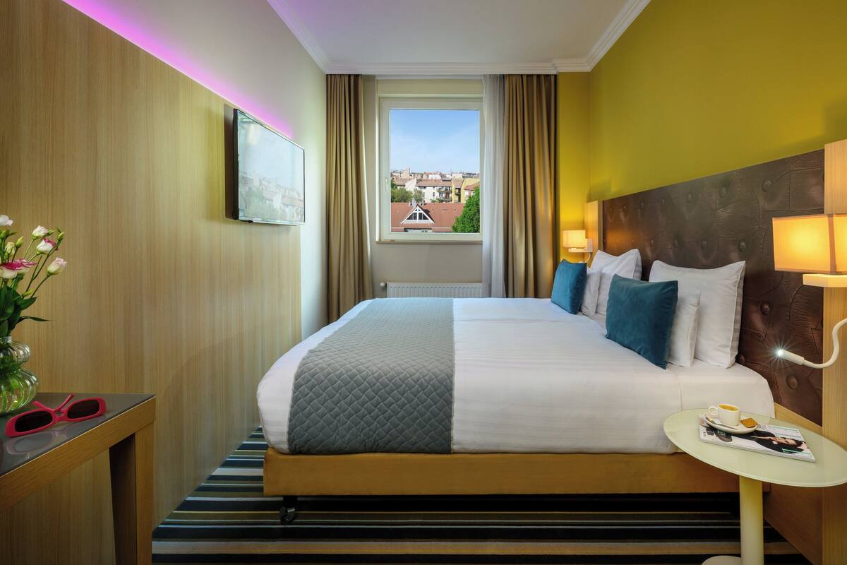 A hotel room with a large bed, a bedside table with a lamp, a window with curtains, and a view of buildings outside.