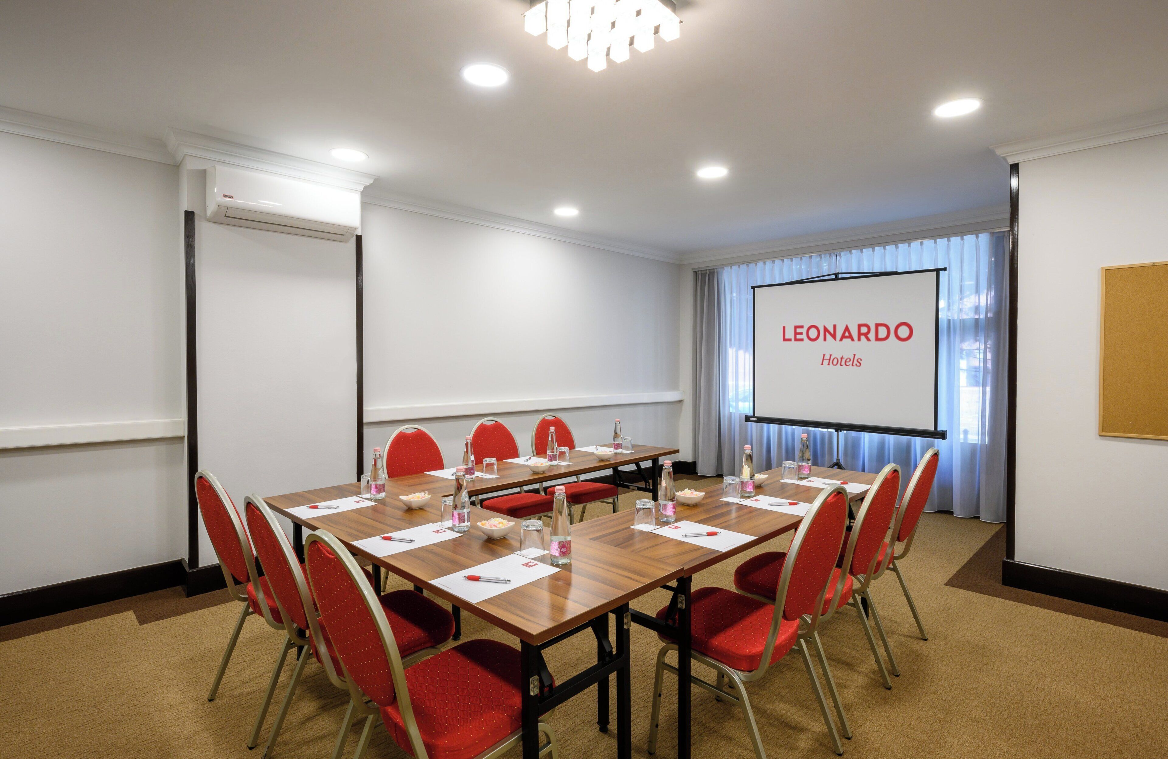 Una sala de conferencias con una larga mesa, sillas rojas, una pantalla de proyector y una pizarra.