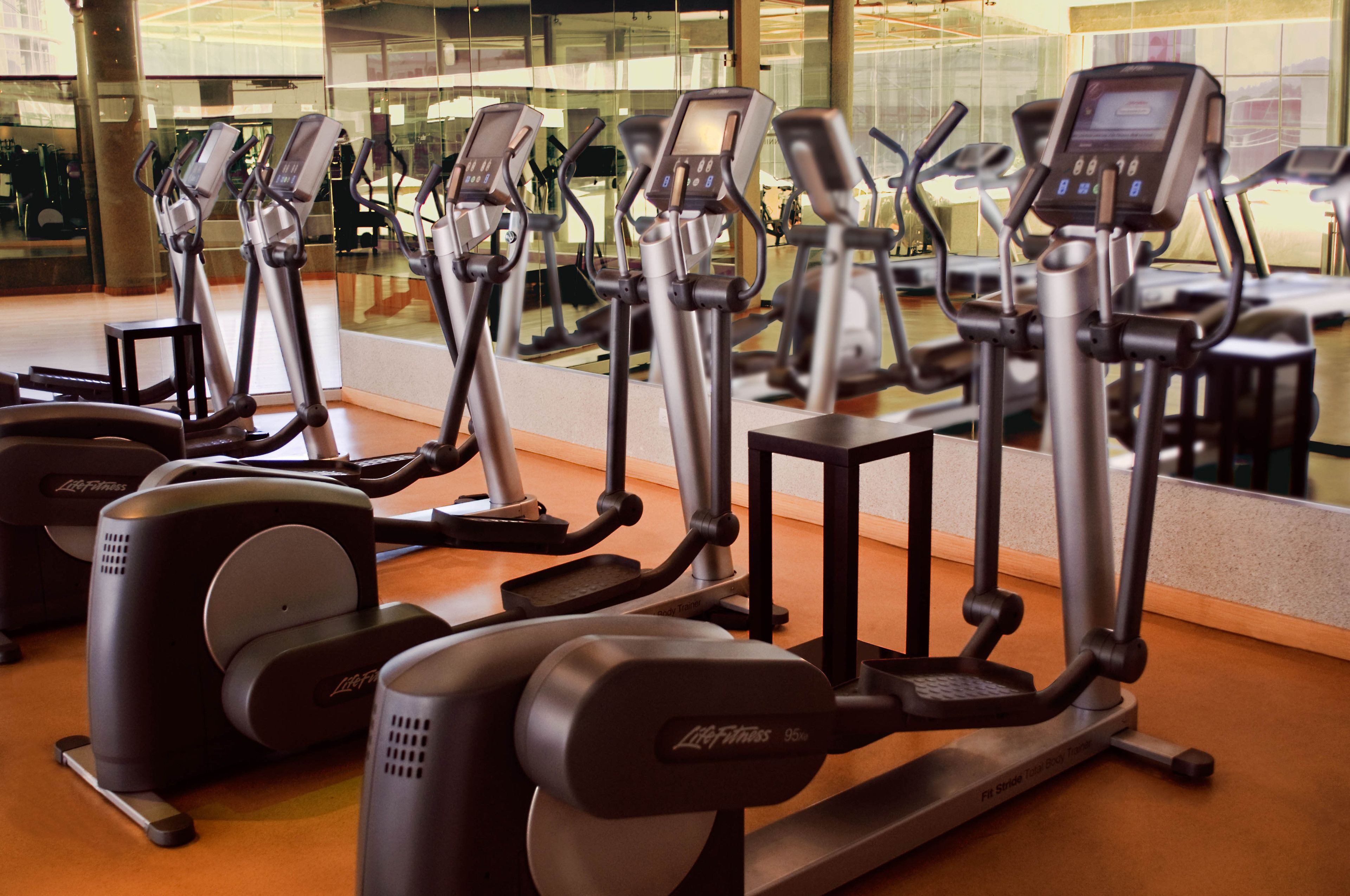 A row of stationary bikes in a gym with a large mirror reflecting the equipment and a glass wall in the background.