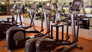A row of stationary bikes in a gym with a large mirror reflecting the equipment and a glass wall in the background.