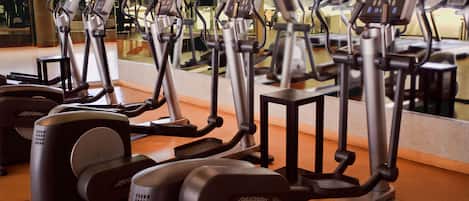 A row of stationary bikes in a gym with a large mirror reflecting the equipment and a glass wall in the background.
