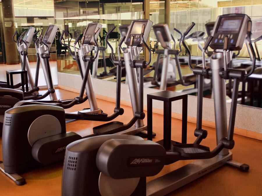 A row of stationary bikes in a gym with a large mirror reflecting the equipment and a glass wall in the background.