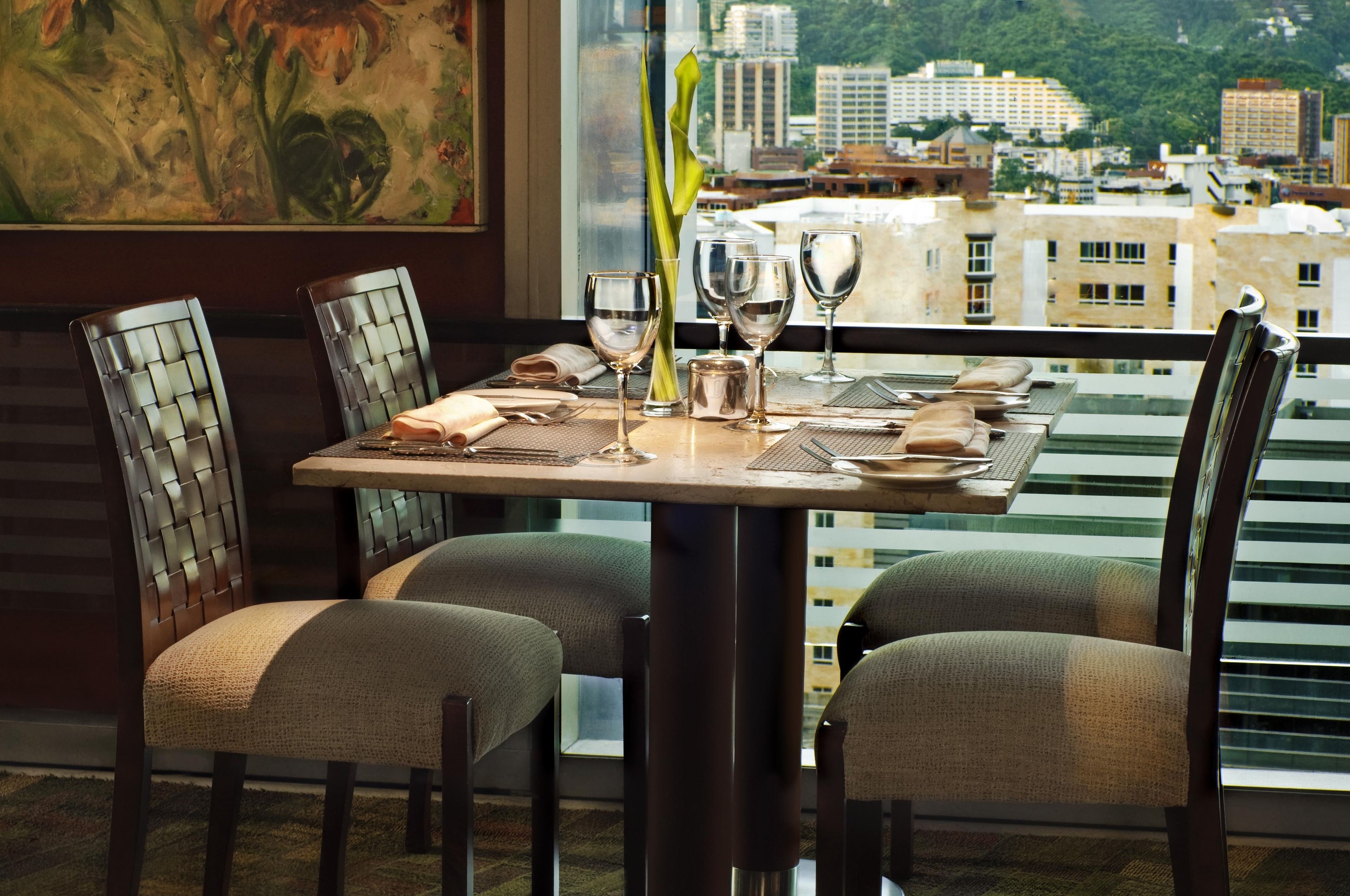A dining table set for four with chairs, a vase of flowers, and a city view through large windows.