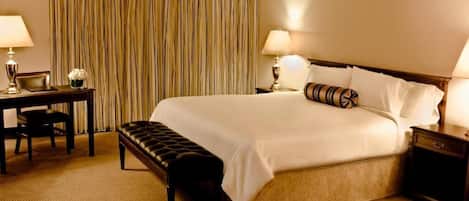 A hotel room with a large bed, two bedside tables with lamps, a desk with a chair, and a bench.