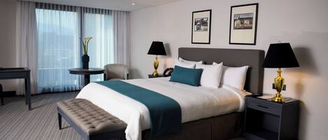 A modern hotel room with a large bed, two bedside tables with lamps, a bench, a desk, and a chair.