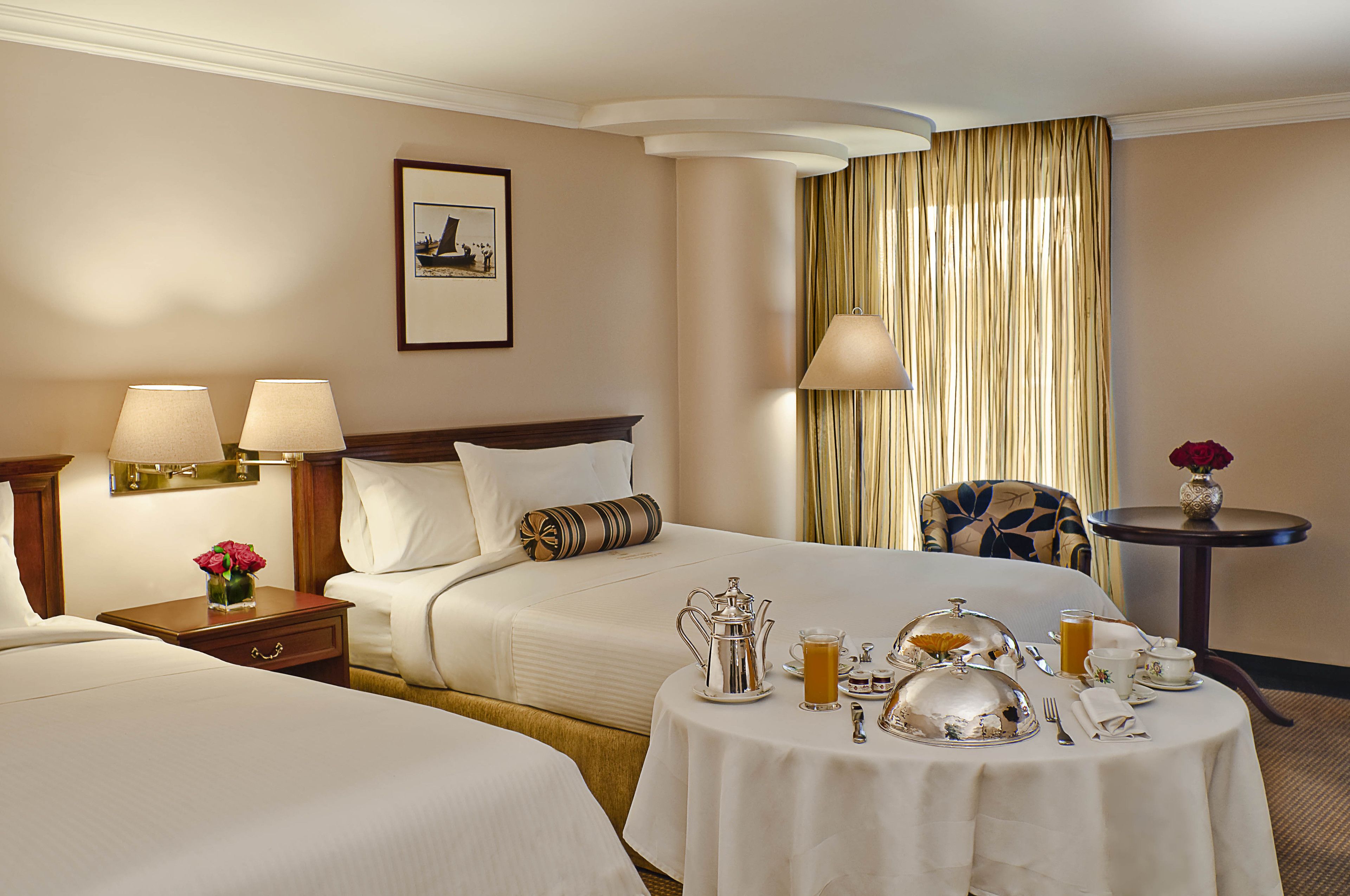 A hotel room with two beds, a dining table set with breakfast, a vase with flowers, and a framed picture on the wall.