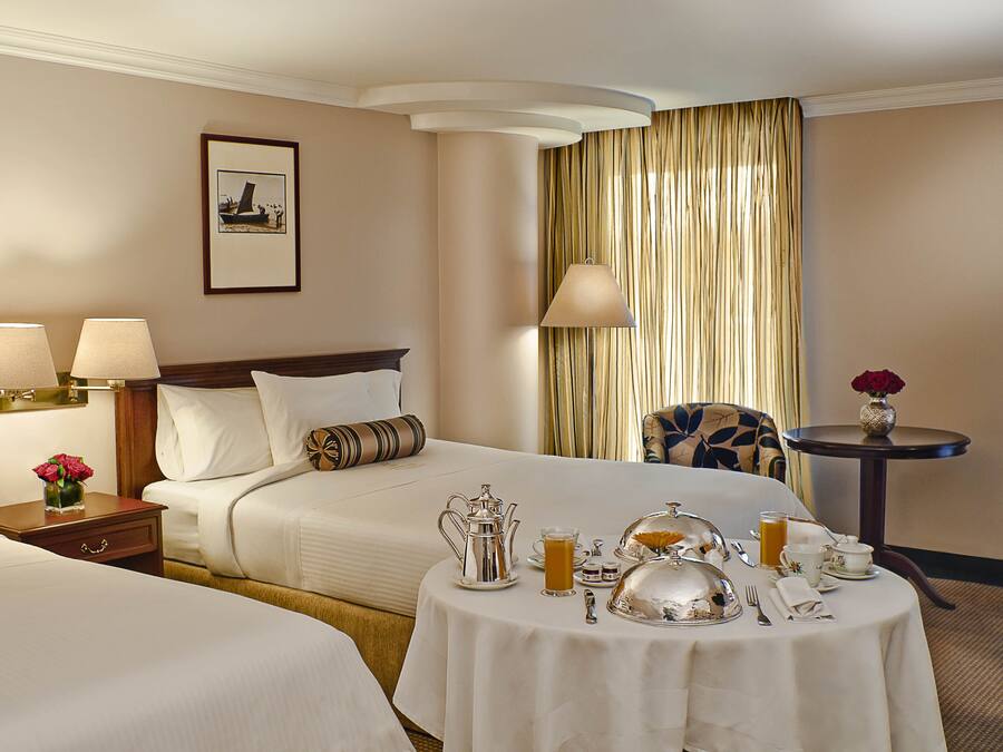 A hotel room with two beds, a dining table set with breakfast, a vase with flowers, and a framed picture on the wall.