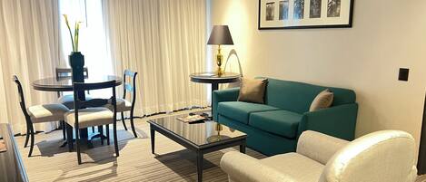 Een hotelkamer met een blauwgroene bank, een beige fauteuil, een ronde salontafel, een eettafel met stoelen en een lamp.