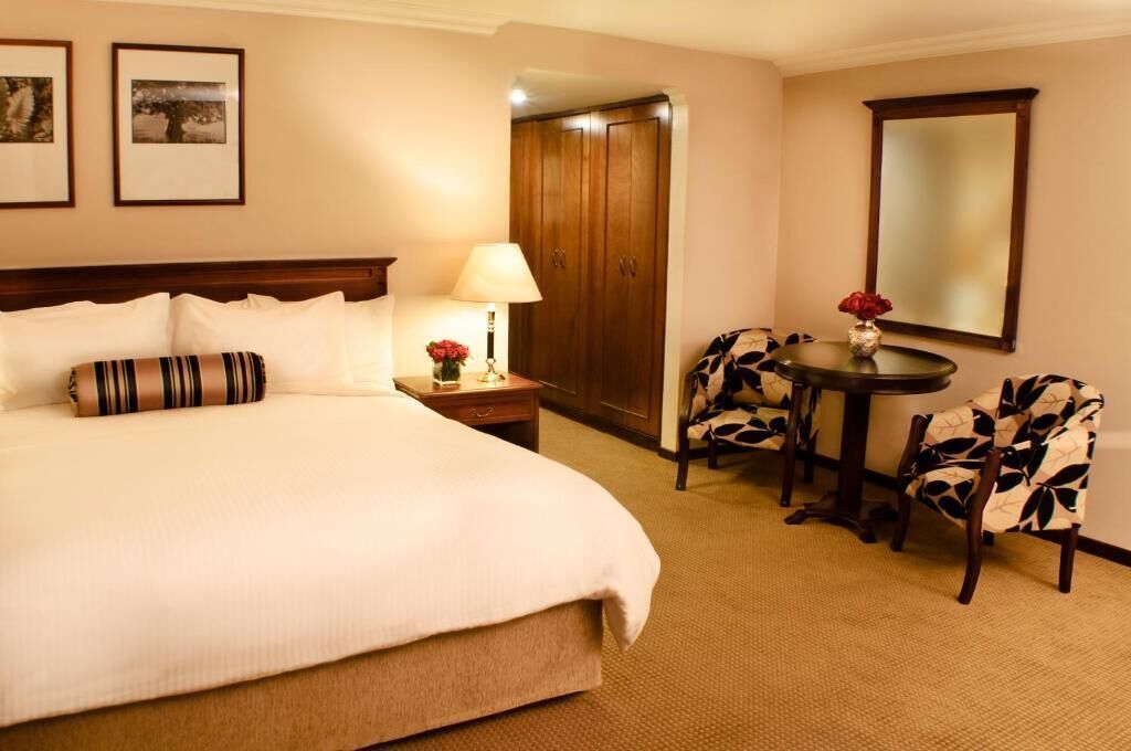 A hotel room with a large bed, a bedside table with a lamp, a small round table with chairs, and a mirror on the wall.