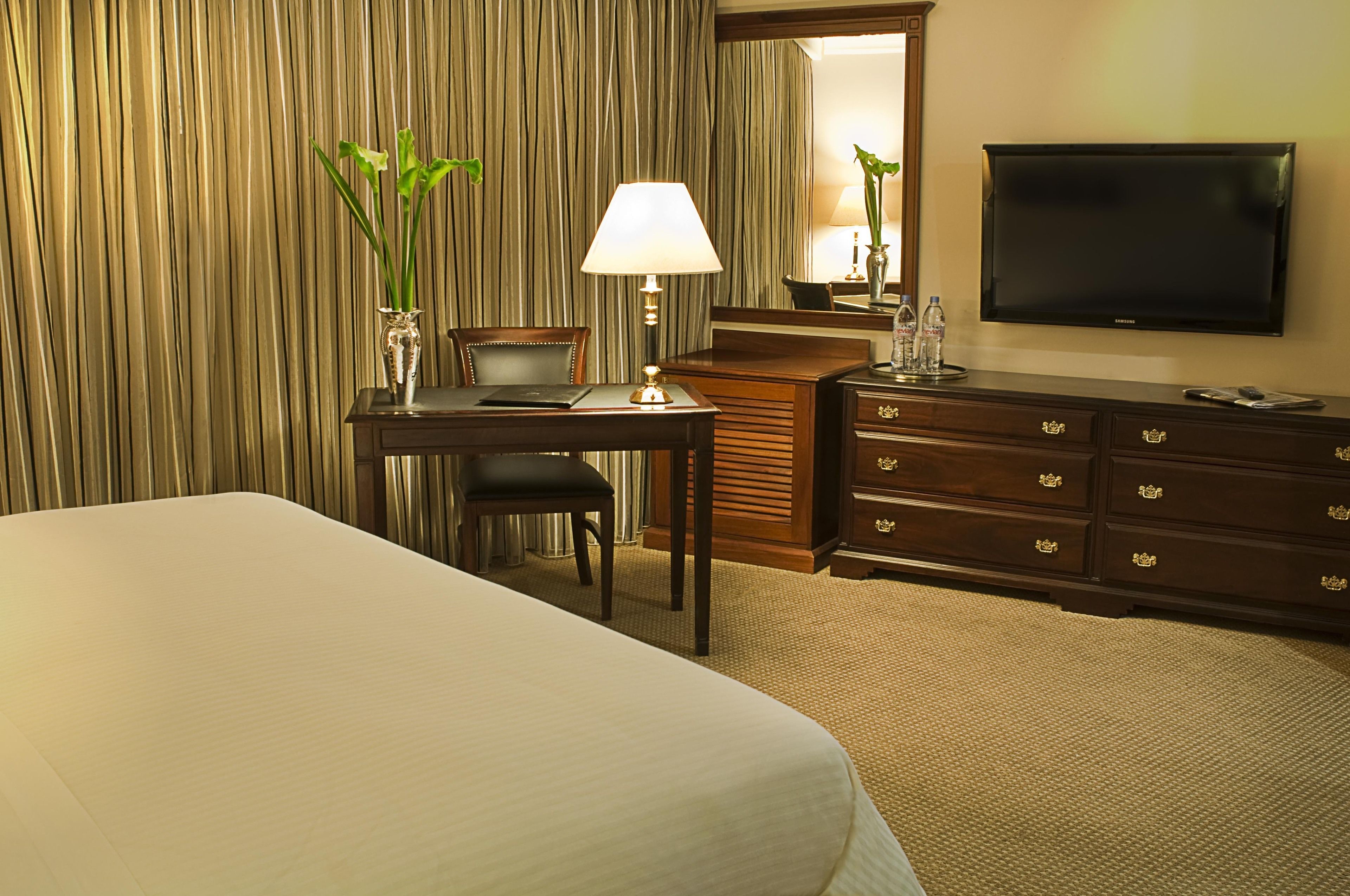 Een hotelkamer met een bed, bureau, stoel, lamp, dressoir, televisie en een spiegel.