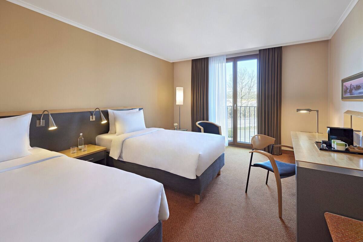 Ein Hotelzimmer mit zwei Betten, einem Schreibtisch, einem Stuhl und einem großen Fenster mit Vorhängen.