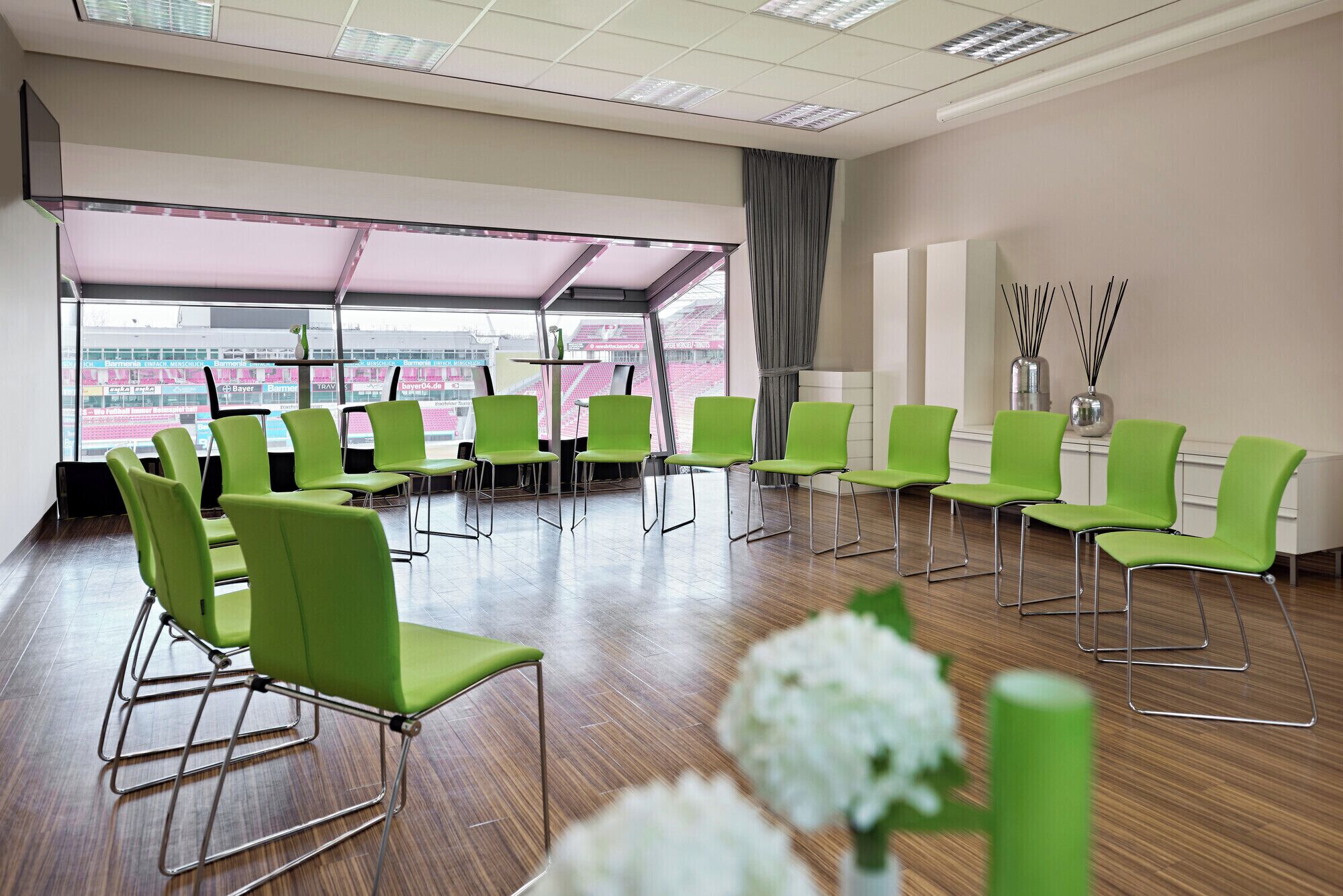 A conference room with green chairs arranged in rows, large windows with a view of a stadium, and a modern ceiling with recessed lighting.