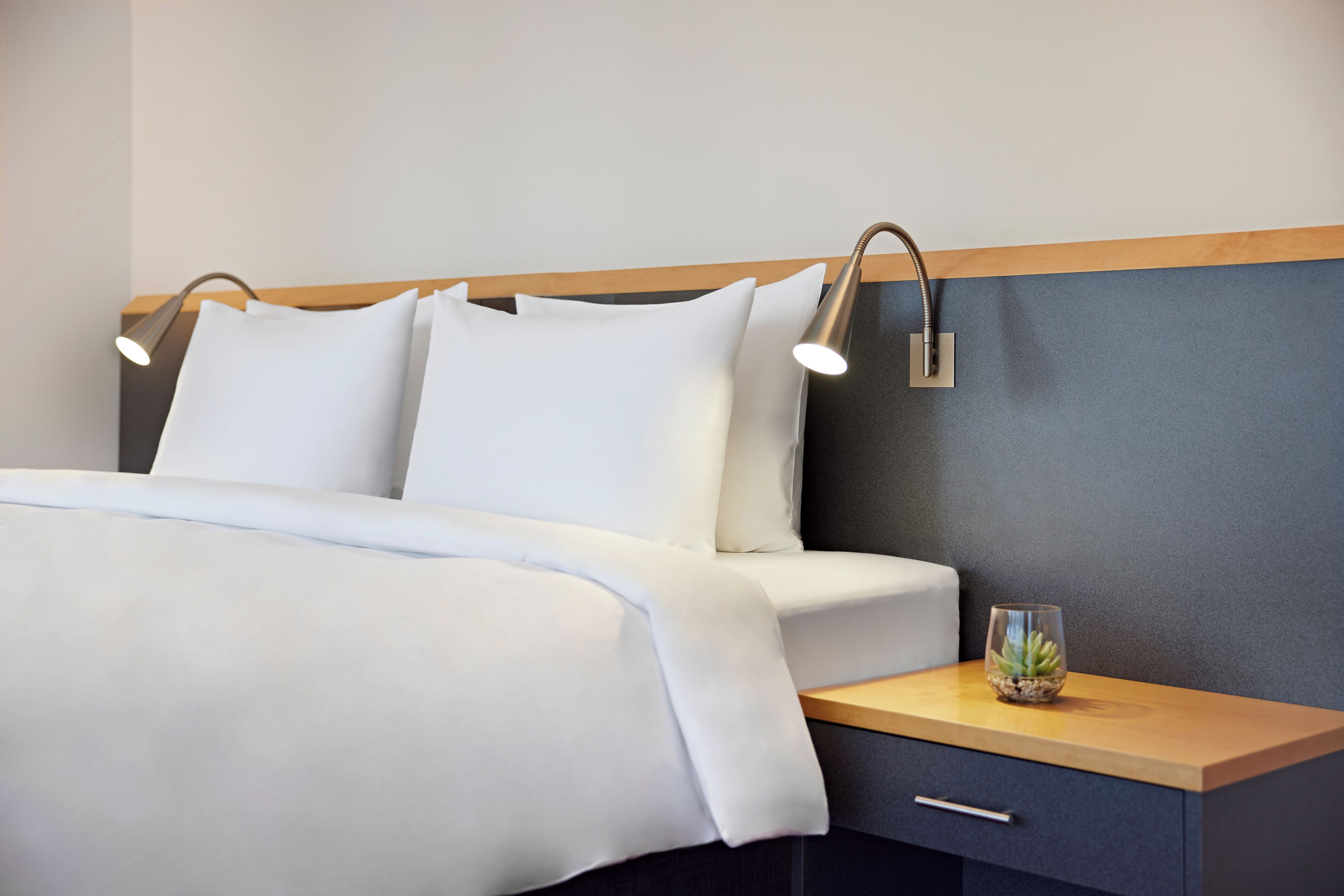 A neatly made bed with white linens, two bedside lamps, a small glass vase with a succulent, and a wooden bedside table with a drawer.