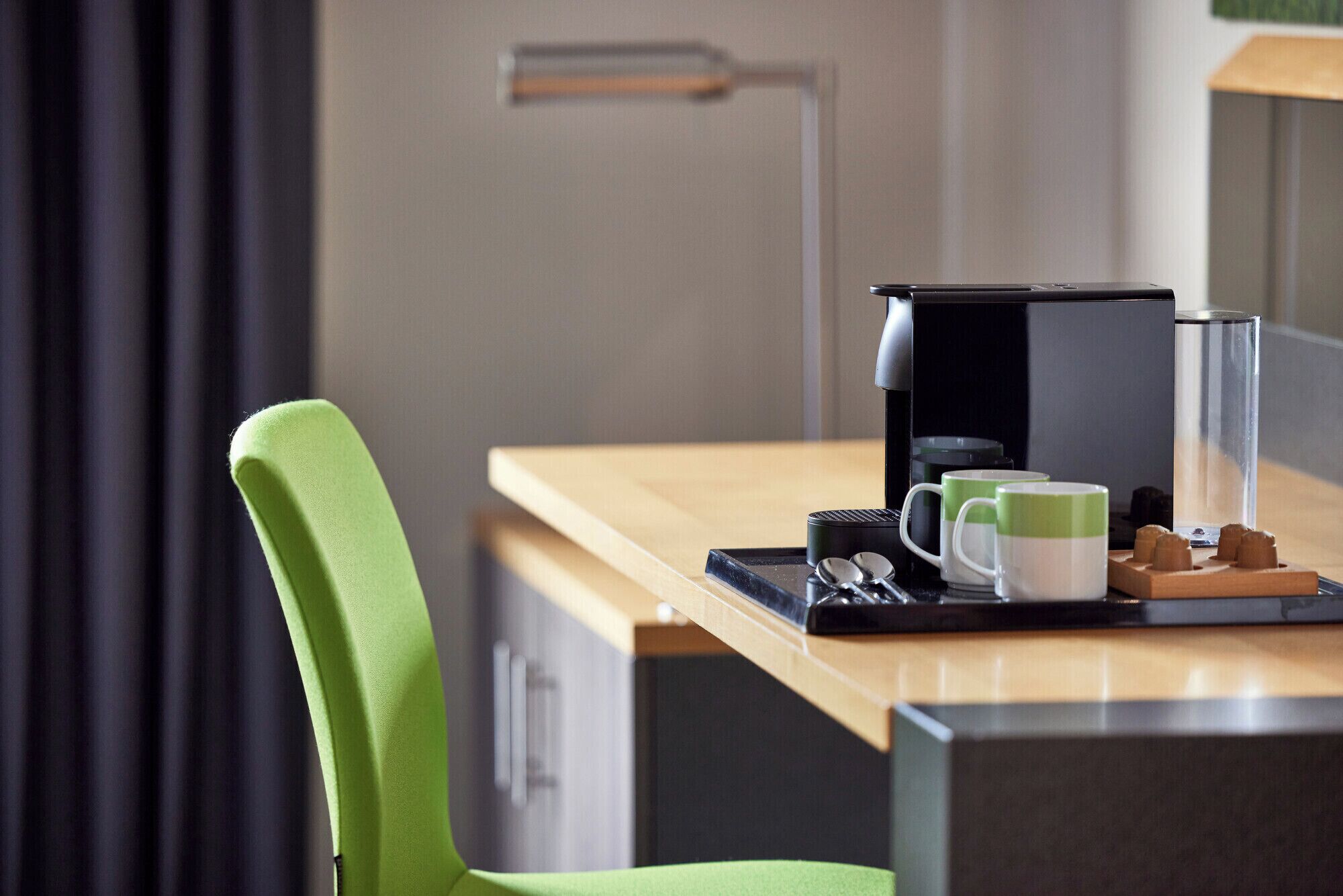 A modern hotel room with a wooden desk, a green chair, a coffee maker, and a tray with mugs and snacks.