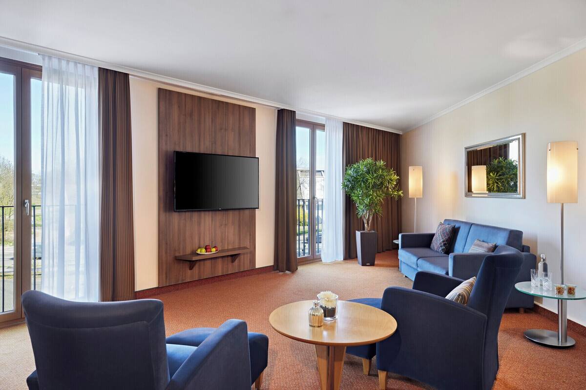 Ein Hotelzimmer mit Flachbildfernseher an der Wand, einer Couch, zwei Sesseln, einem kleinen runden Tisch mit einer Vase und einer Topfpflanze.