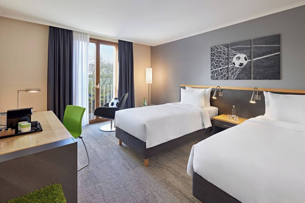 Ein Hotelzimmer mit zwei Betten, einem Schreibtisch mit Lampe, einem Sessel und Blick auf Bäume durch das Fenster.