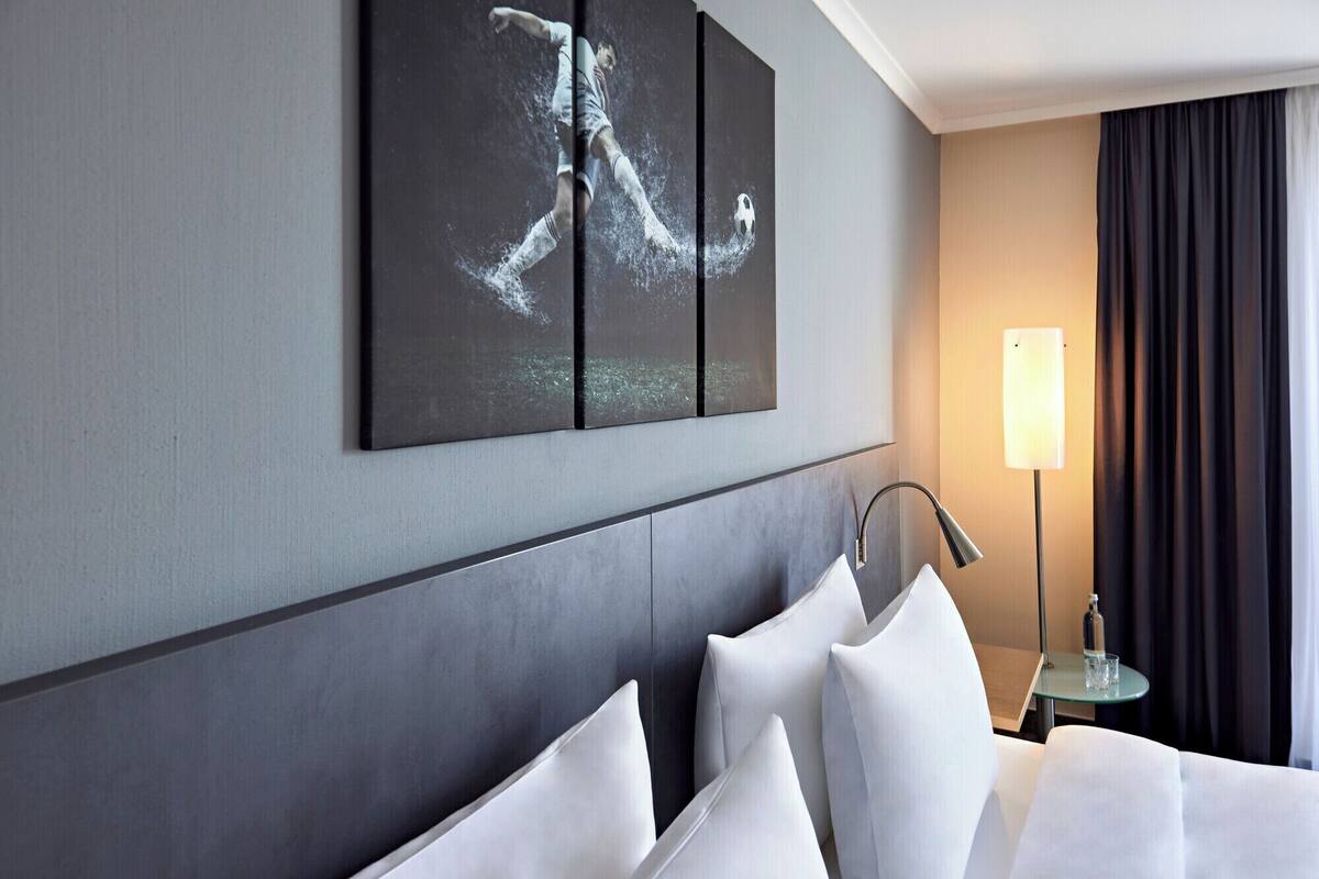 Ein modernes Hotelzimmer mit einem großen Bett, einem Wandbild, einer Nachttischlampe und Blick auf den Nachthimmel durch ein Fenster.