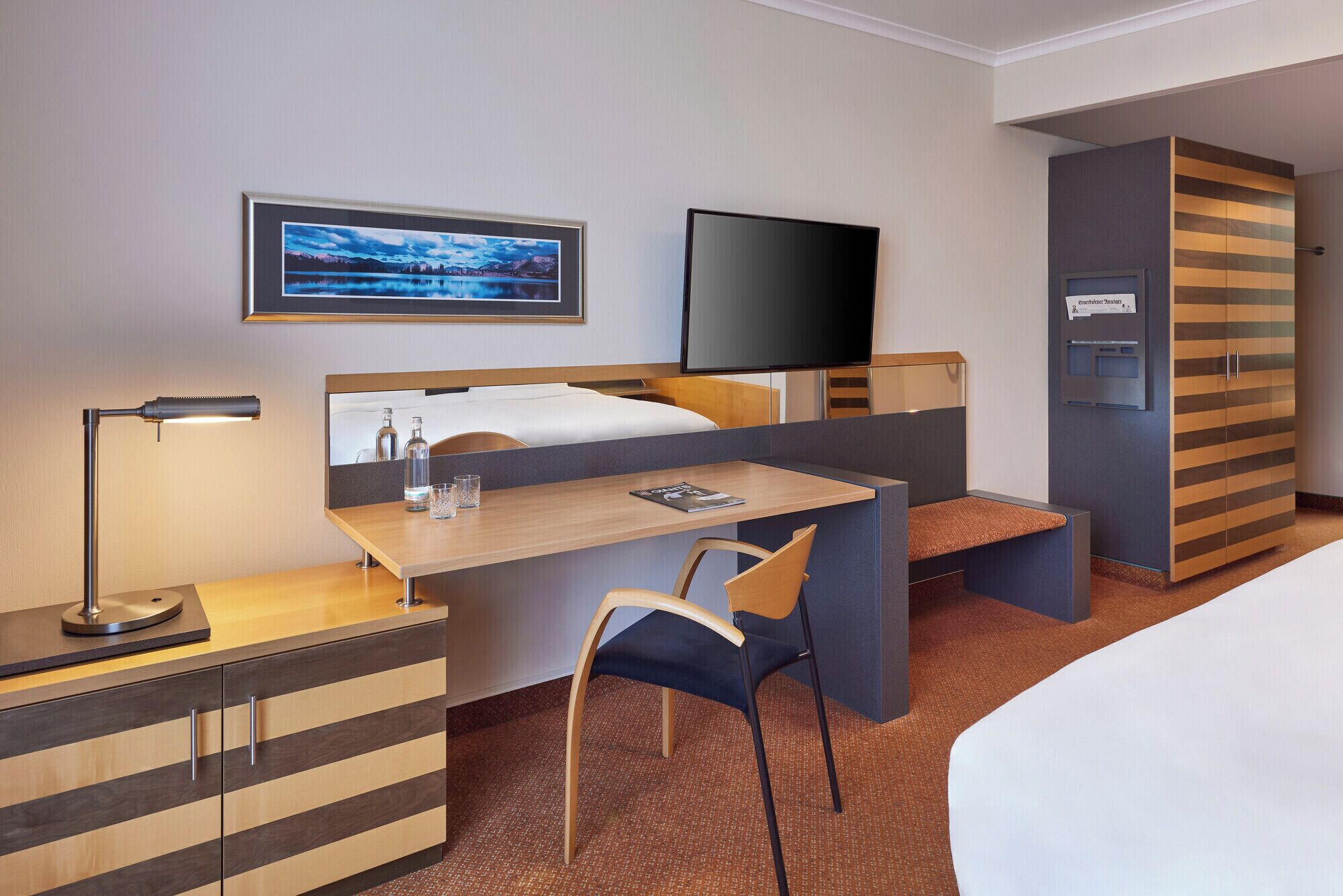 A hotel room with a bed, a desk, a chair, a TV, and a minibar.