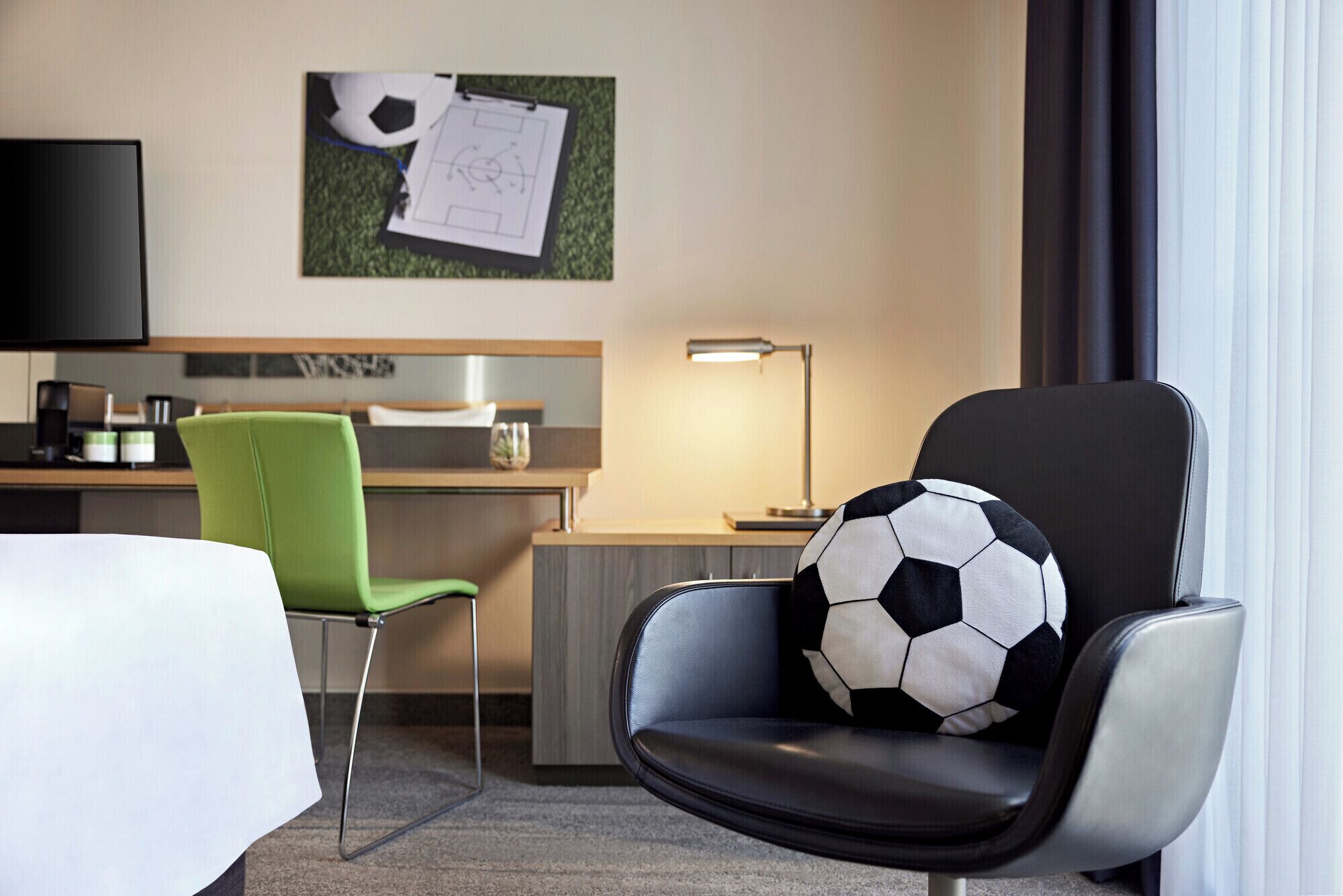 A hotel room with a single bed, a green chair, a black leather armchair with a soccer ball cushion, a desk with a lamp, and a wall-mounted TV.