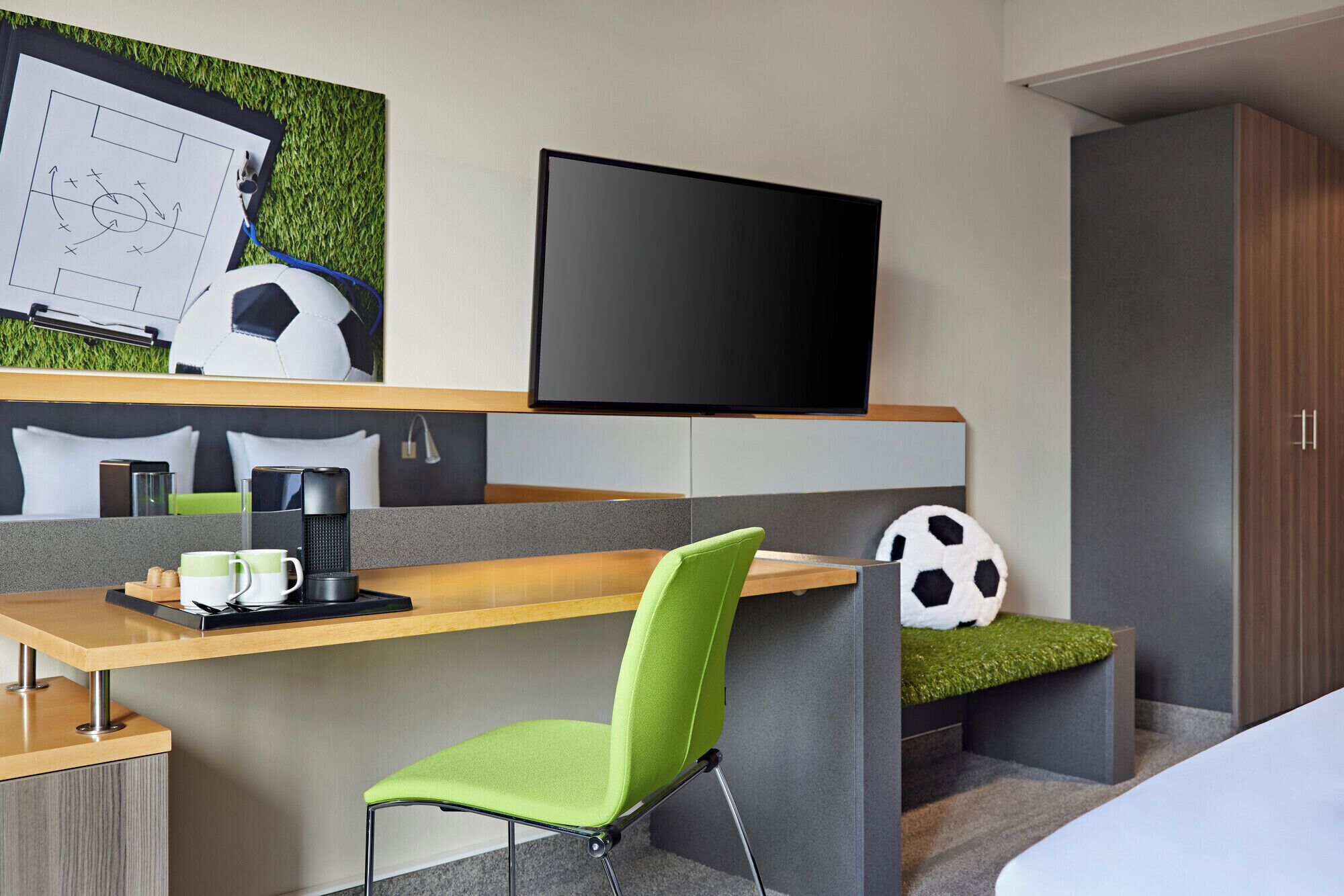 A modern hotel room with a wooden desk, a green cushioned chair, a flat-screen TV, a soccer ball, and a bed with a green grass-like cover.
