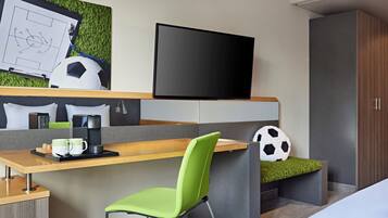 Een moderne hotelkamer met een houten bureau, een groene fauteuil, een flatscreen televisie, een voetbal en een bed met een deken die op gras lijkt.