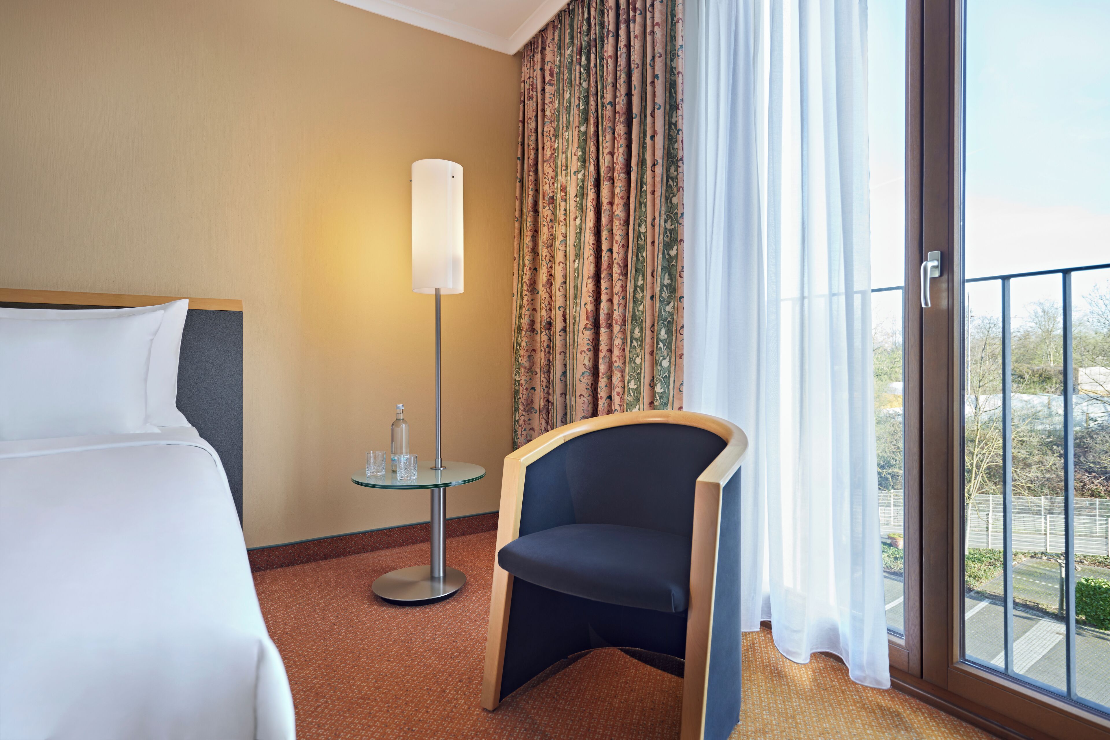 A hotel room with a bed, a chair, a table with a lamp, and a view through a window.