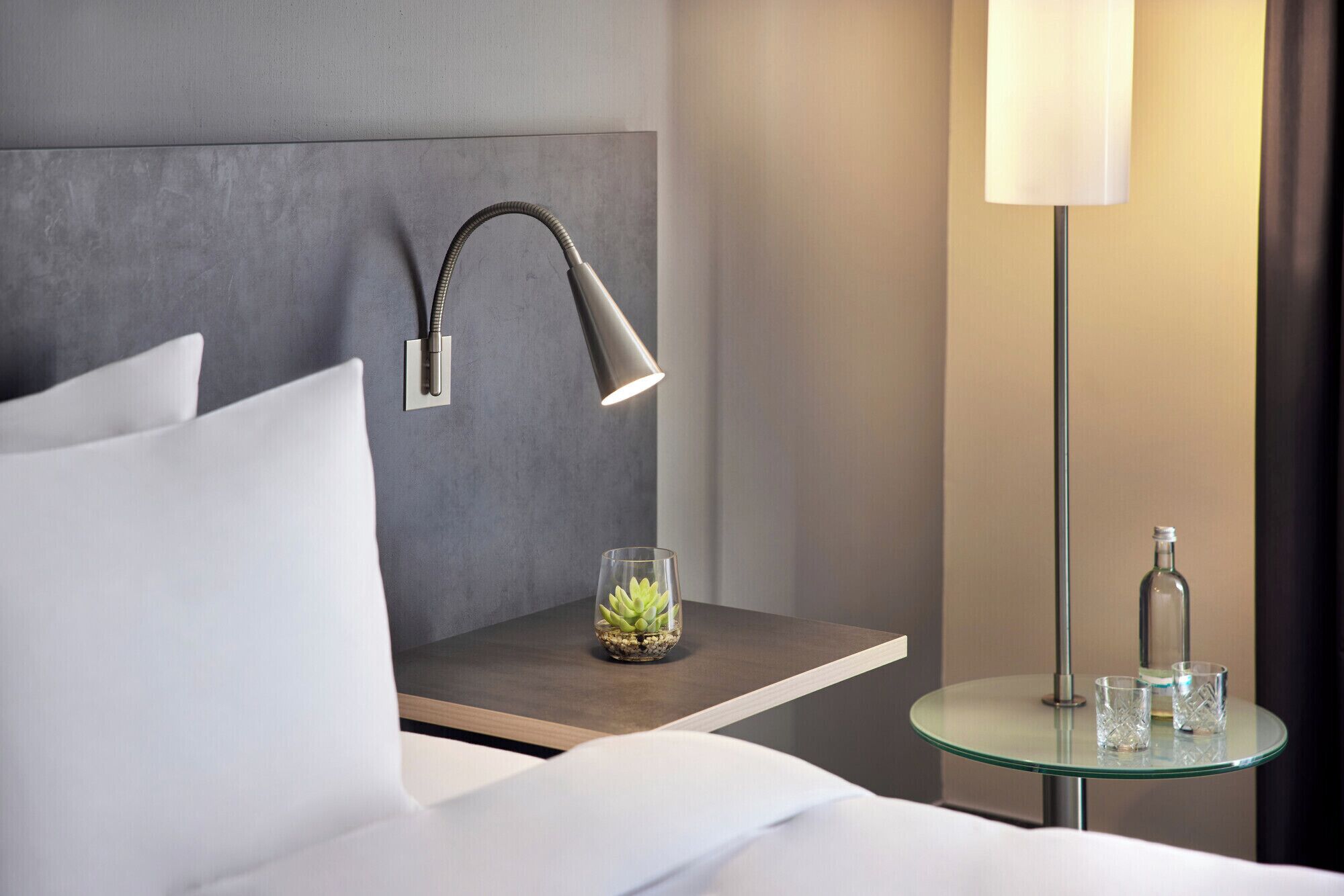 A neatly made bed with white linens, a bedside table with a lamp, a small glass table with a glass and a bottle, and a wall-mounted lamp.