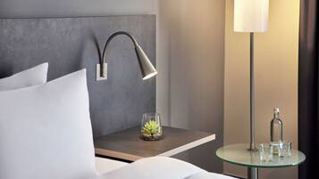 Ein ordentlich bezogenes Bett mit weißen Leinen, ein Nachttisch mit Lampe, ein kleiner Glastisch mit Glas und Flasche sowie eine Wandlampe.