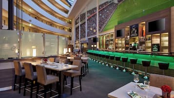 Un moderno vestíbulo de hotel con techo alto, una zona de bar con taburetes y un área de comedor con mesas y sillas.