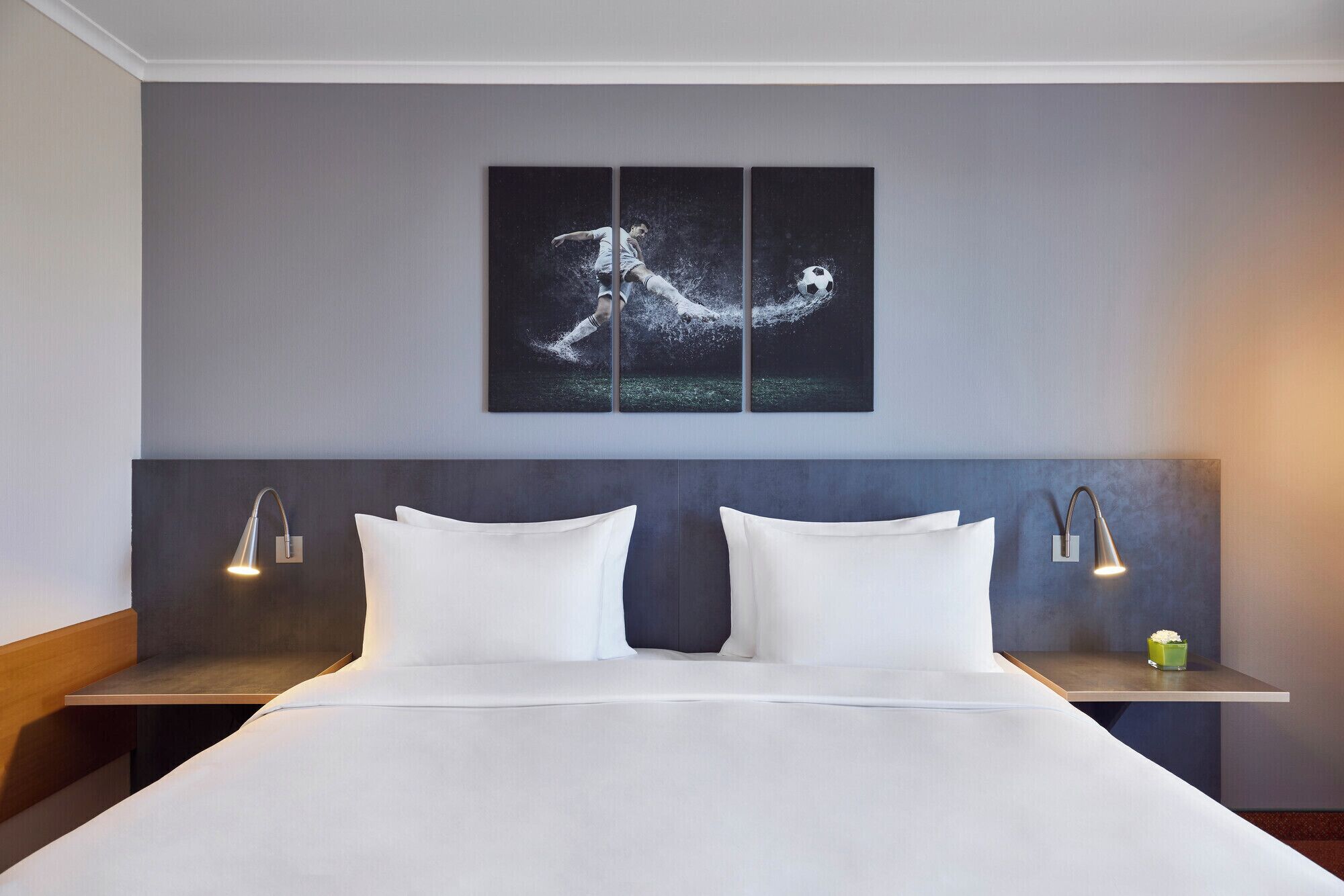 A modern hotel room with a large bed, two bedside tables, a headboard, and a wall-mounted artwork depicting a soccer player.