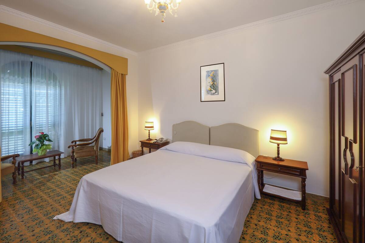 Una habitación de hotel con una cama grande, dos mesitas de noche con lámparas, una silla, una pequeña mesa con un florero y una ventana con cortinas.