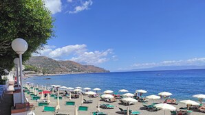 A beach with rows of sun loungers and umbrellas, a clear blue sky, and a view of the sea and distant hills. - Hotel Lido Mediterranee (Taormina)