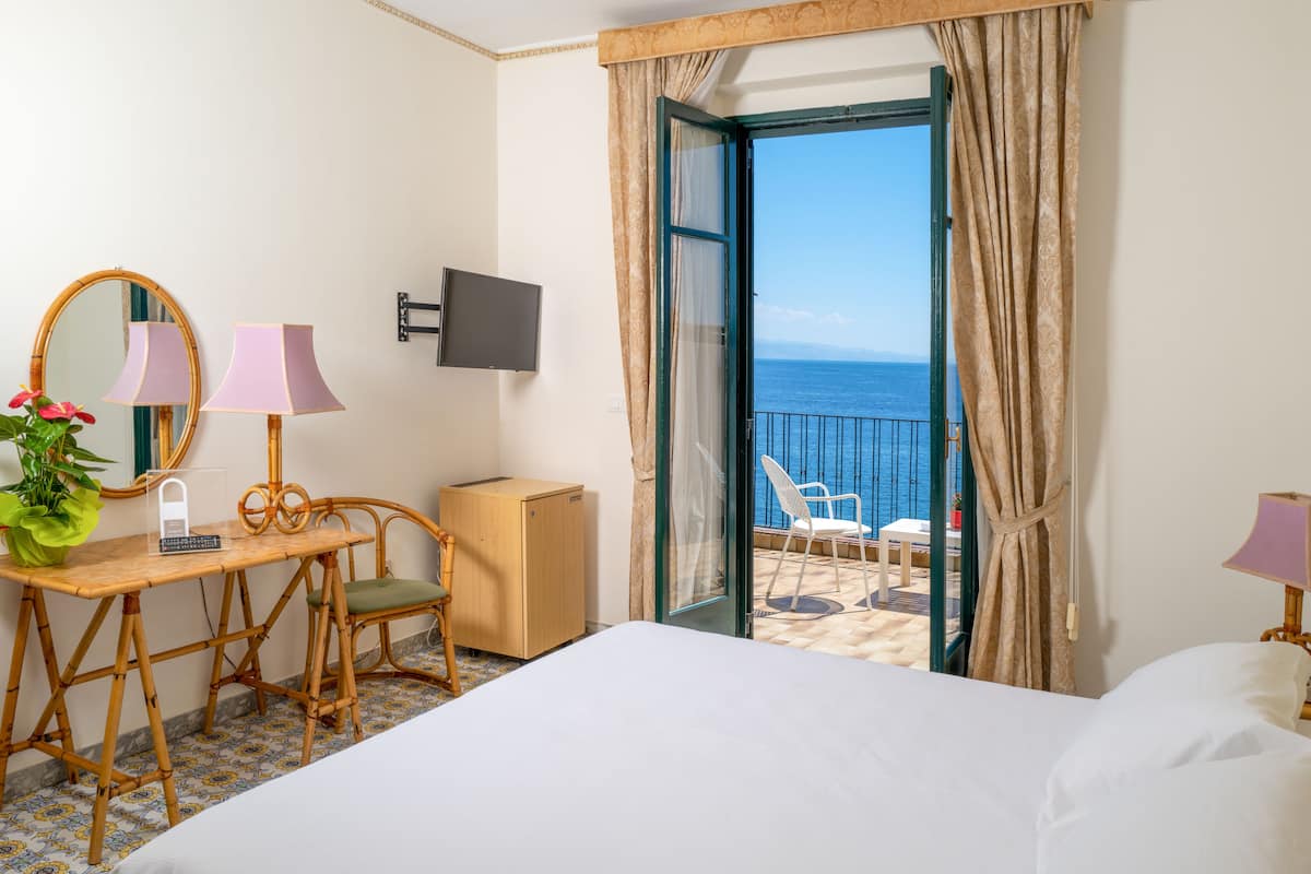 Habitación de hotel con cama, escritorio con lámpara, silla, pequeño armario, televisor y balcón con vista al mar.