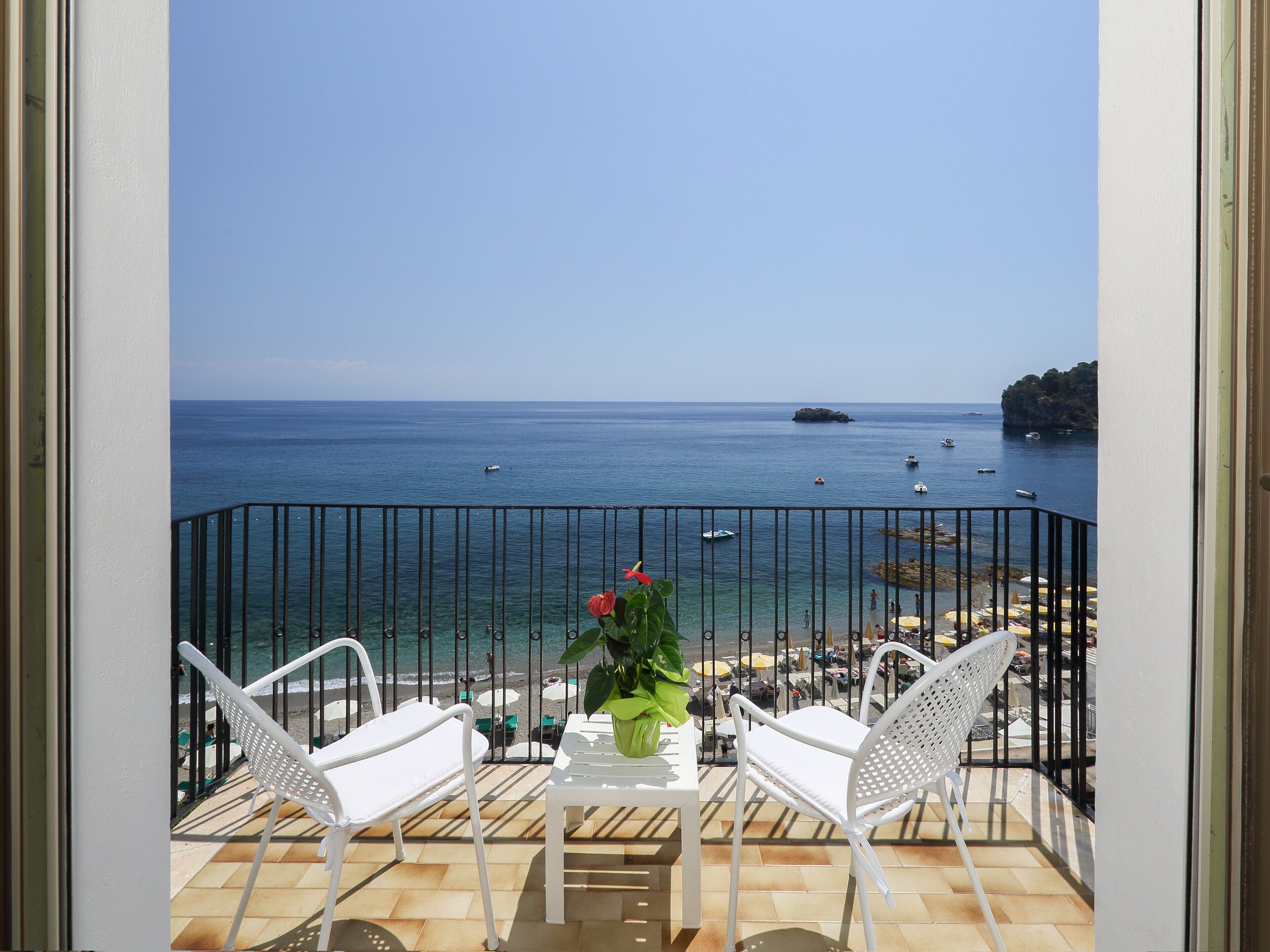A balcony with white chairs and a table, overlooking a beach with boats and a rocky outcrop.