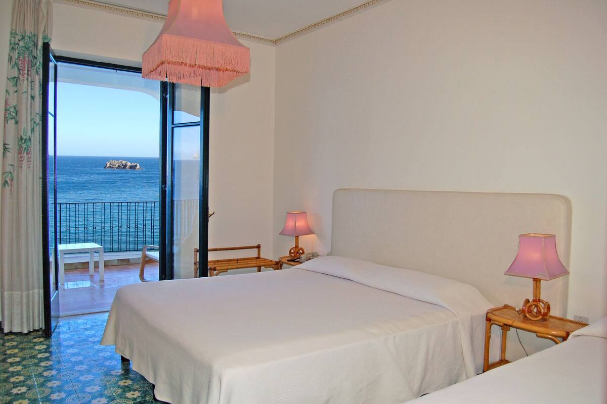 Habitación de hotel con una cama grande, dos mesitas de noche con lámparas, una silla y vistas al mar a través de una puerta abierta.