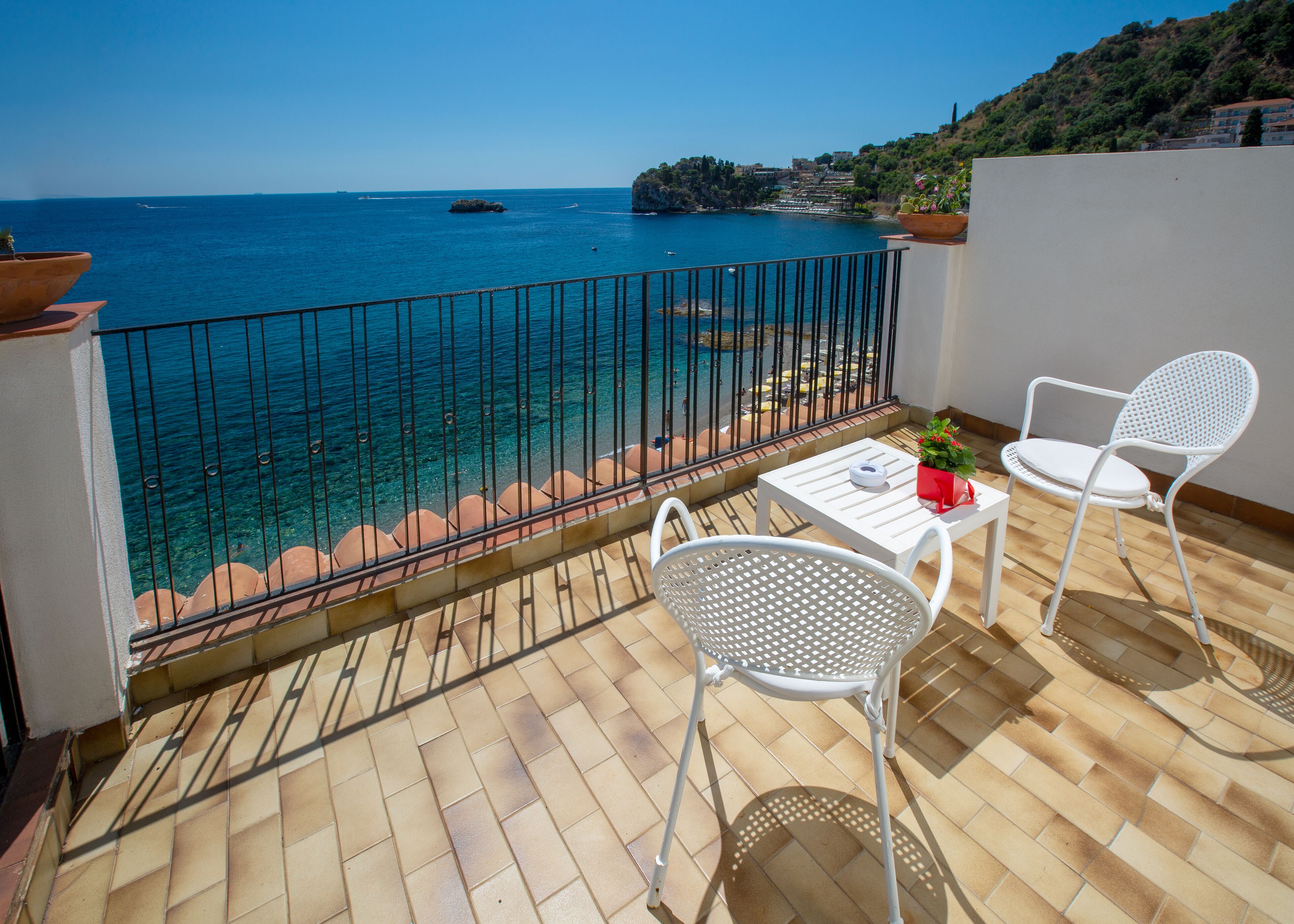 A balcony with a view of the sea, two white chairs, a small table, and a potted plant.