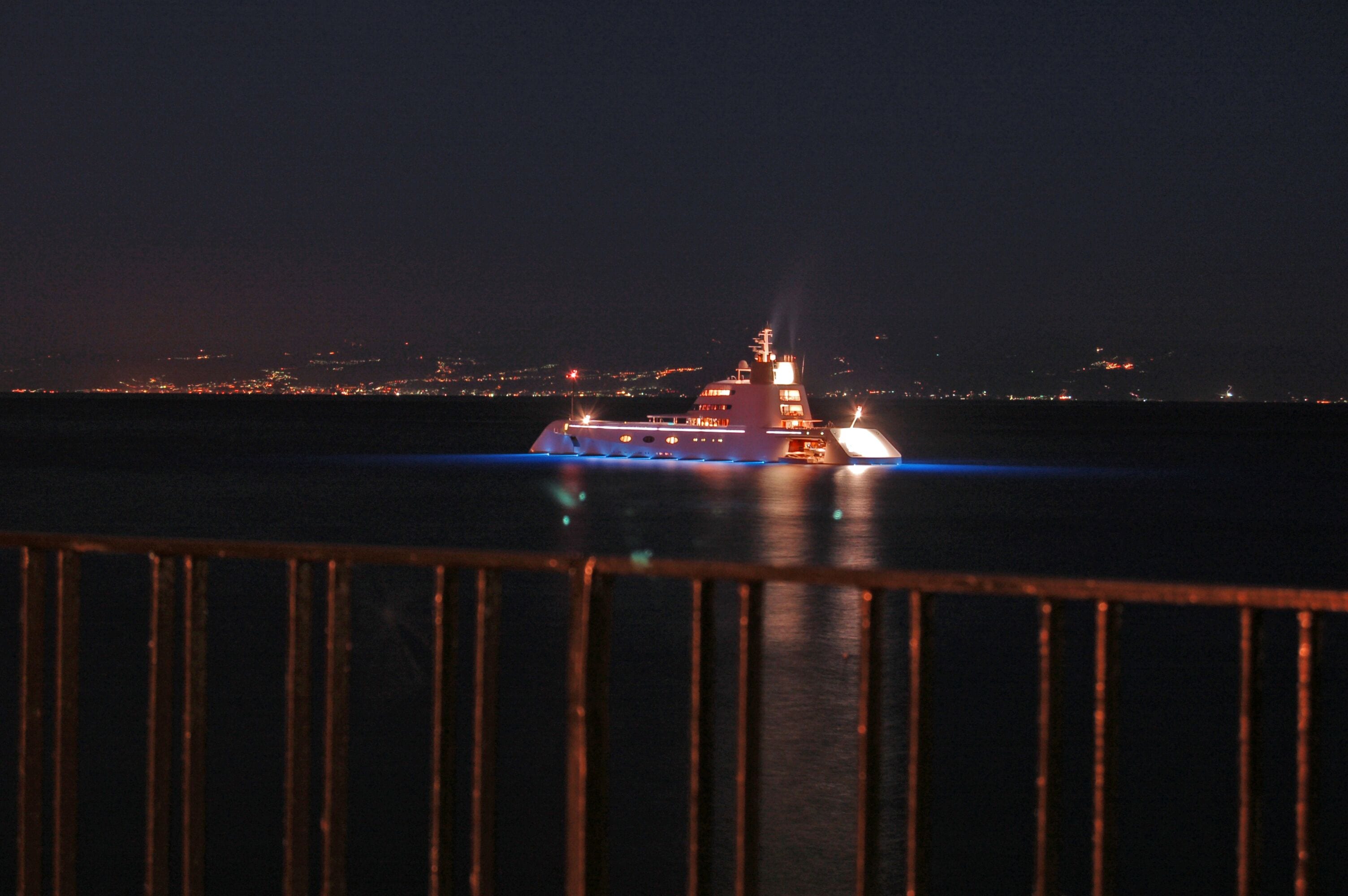 A ship at night with lights on, a railing in the foreground, and a cityscape in the background.