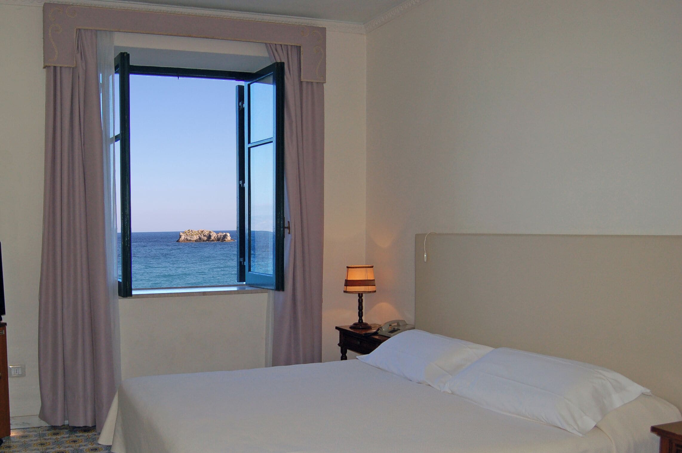A hotel room with a bed, a nightstand with a lamp, and a window with curtains.