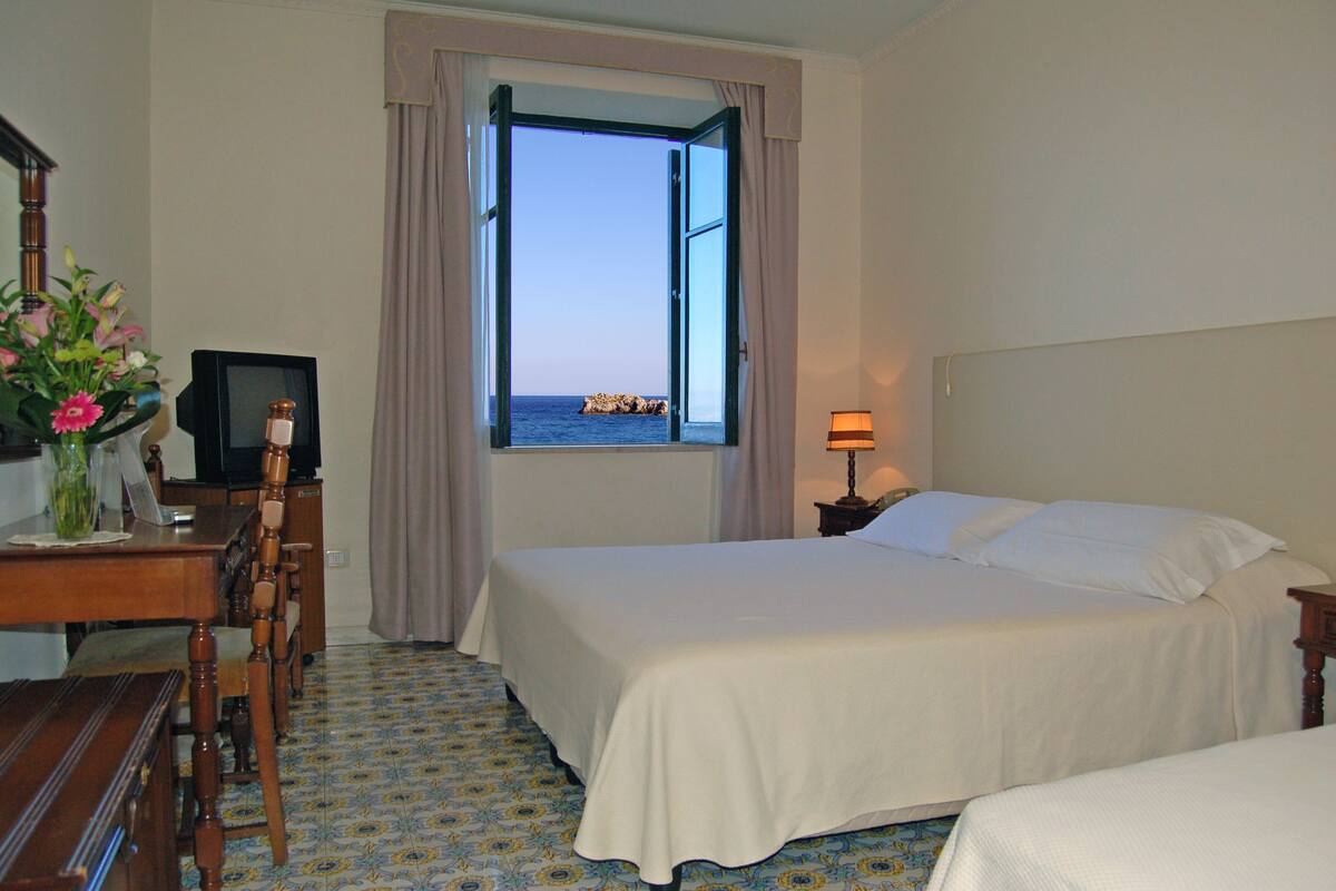 Habitación de hotel con cama, escritorio con televisor, lámpara y vista al mar a través de una ventana abierta.