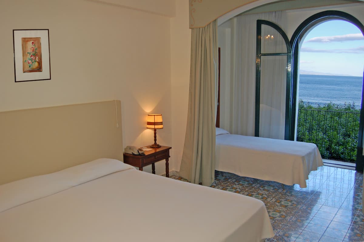 Habitación de hotel con dos camas, una mesita de noche con lámpara, un cuadro en la pared y un balcón con vista al mar.