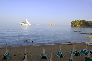 A beach with lounge chairs and umbrellas, a yacht in the water, and a rocky cliff with buildings.