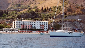 A coastal building with multiple balconies overlooking the sea, a sailboat docked nearby, and a beach with umbrellas and people. - Hotel Lido Mediterranee (Taormina)