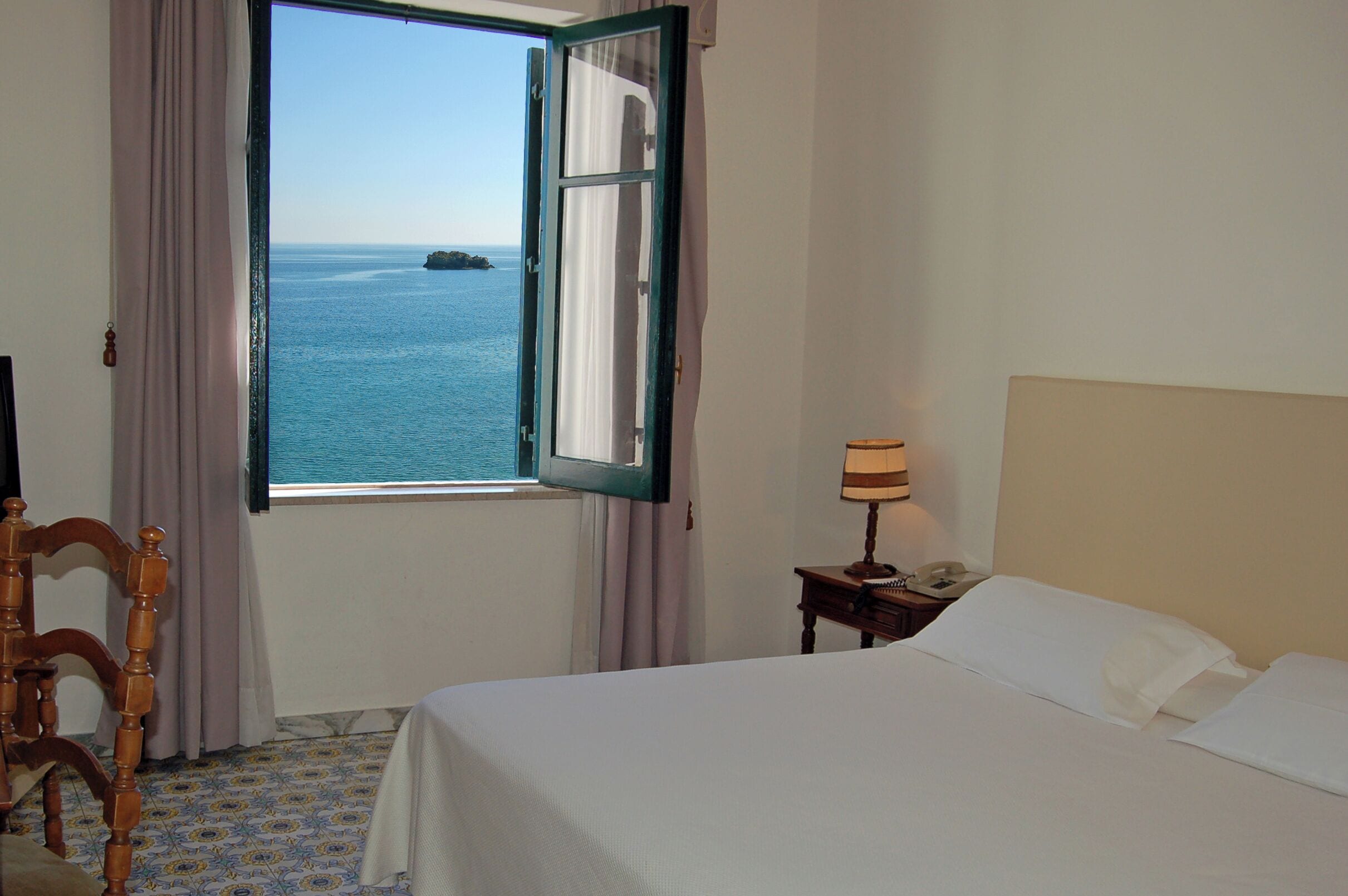A hotel room with a bed, a nightstand, a lamp, a chair, and a window with a view of the sea.